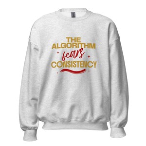 May include: A light gray crewneck sweatshirt featuring the gold text THE ALGORITHM and CONSISTENCY with the red script word fears in the center. The design includes small red star accents and a red brushstroke underline on the front chest.
