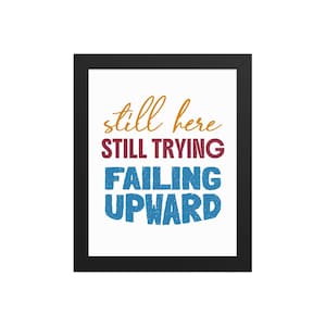 May include: A framed wall art print featuring the text "still here" in orange cursive, "STILL TRYING" in maroon sans-serif capital letters, and "FAILING UPWARD" in blue textured sans-serif capital letters on a plain white background.