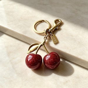 Può includere: Un portachiavi in metallo dorato con due ciliegie rosso scuro lucide e sottili riflessi glitterati. Il ciondolo include un dettaglio a foglia dorata, un anello circolare, un moschettone e una piccola targhetta rettangolare con inciso COACH.