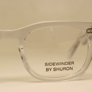 Puede incluir: Gafas con montura de plástico transparente con una etiqueta negra que dice "SIDEWINDER BY SHURON".
