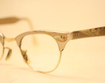 May include: Vintage cat-eye spectacles with a gold and silver frame. The frame has delicate floral engravings on the temples and around the lenses. The lenses are clear, and the overall design is retro.