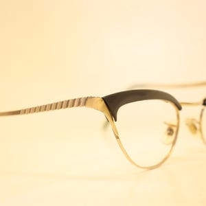 Puede incluir: Gafas vintage de ojo de gato con montura de metal dorado y detalles en negro. Las patillas tienen un diseño a rayas. Las lentes son transparentes. Un estilo retro clásico.