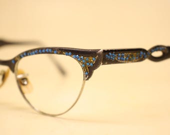 Könnte beinhalten: Nahaufnahme einer Vintage-Brille in Cat-Eye-Form. Die dunklen Rahmen sind mit kleinen blauen und goldenen Verzierungen verziert. Die Gläser sind klar, und die Metallbügel haben ein dekoratives Design.