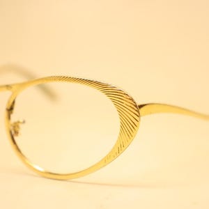 May include: Close-up of gold-colored eyeglasses with oval lenses. The frames have a textured design on the outer edges and temples. The glasses are set against a soft, neutral background.