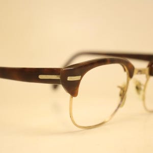 May include: Eyeglasses featuring a brown tortoiseshell pattern on the upper brow bar and temples, complemented by a thin gold-tone metal wire frame along the bottom of the lenses. The design includes small silver-tone rectangular accents on the hinges.