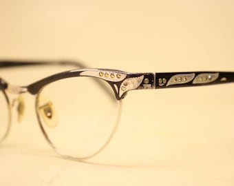 Könnte beinhalten: Vintage Cat-Eye-Brille mit schwarzem Rahmen und silbernen Akzenten. Die Fassungen haben ein dekoratives Design an den Bügeln und oberen Rändern. Die Gläser sind klar, und die Brille hat eine Retro-Ästhetik.