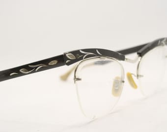 May include: Vintage cat-eye glasses with a black frame and silver accents. The frame has a floral design etched into it.