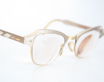 May include: A pair of vintage cat-eye glasses with a light brown frame and gold accents. The frame has a delicate floral design.
