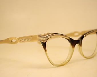 May include: Vintage cat-eye spectacles with a gradient brown and clear frame. The arms and temples are a light beige colour with leaf and swirl detailing. The glasses have a classic, retro style.