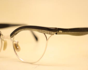 May include: Vintage cat-eye style spectacles. The frames are made of silver metal and black plastic. The upper rims feature a decorative pattern. The lenses are clear.