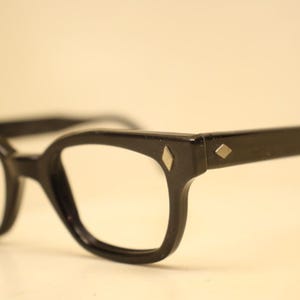 May include: Black eyeglasses with a thick frame and a distinct diamond-shaped silver metal inlay on the outer corners of the front frame and on the temples. The glasses feature a classic rectangular lens shape with rounded edges and sturdy arms.