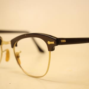 Puede incluir: Gafas vintage con ceja negra y montura de metal dorado. Las gafas tienen un diseño clásico y retro con una parte superior curvada y detalles rectangulares. Las lentes son transparentes.