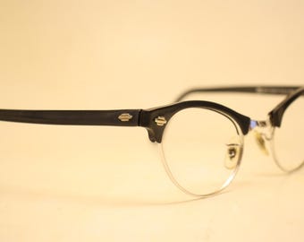 May include: Vintage spectacles with a black frame and clear lenses. The glasses have a half-rim design with a silver-coloured metal frame on the bottom half. The temples are black and feature small gold-coloured accents. The overall style is retro.