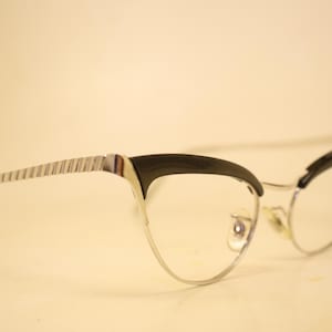 Puede incluir: Gafas de estilo ojo de gato con montura plateada y bordes superiores negros. Las patillas tienen un diseño a rayas. Las lentes son transparentes. Las gafas están sobre un fondo claro.