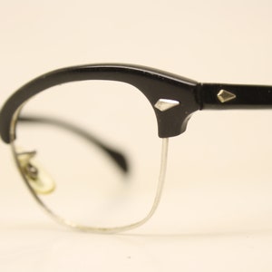 Op de afbeelding: Zwarte bril met een plastic montuur, zilveren metalen pootjes en diamantvormige accenten op de pootjes.