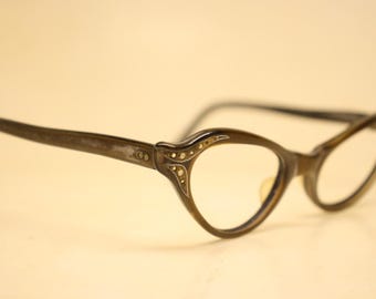 May include: Vintage cat-eye spectacles with a dark brown frame. The frames feature decorative detailing with small, gold-coloured accents near the hinges. The lenses are clear, and the overall style is retro.