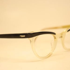 Puede incluir: Gafas con montura transparente y detalles negros en los bordes superiores y las patillas. Las gafas tienen forma de ojo de gato. Las lentes son transparentes, sobre un fondo suave y neutro.