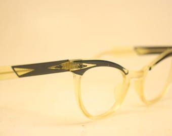 May include: Vintage cat-eye spectacles with a clear frame and black and gold detailing. The arms are a light yellow colour. The glasses have a retro style.