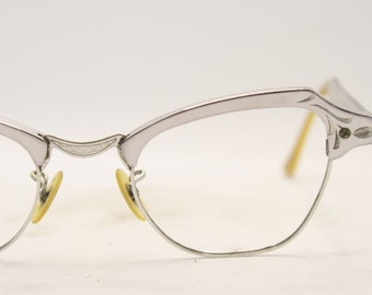 May include: Silver-toned eyeglasses with a cat-eye design. The frames have decorative detailing on the upper rims and temples. The lenses are clear, and the nose pads are yellow. A vintage style.