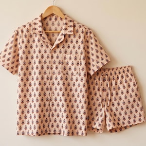 May include: A short-sleeve button-down shirt and matching shorts set in a light peach color featuring a repeating dark blue and red floral block print pattern. The shirt has a notched collar, a single chest pocket, and a front button closure.