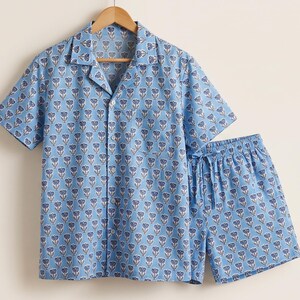 May include: A light blue short-sleeve button-down shirt and matching shorts set featuring a repeating dark blue and white floral pattern. The shirt includes a notched collar, a single chest pocket, and white buttons, while the shorts have a drawstring.