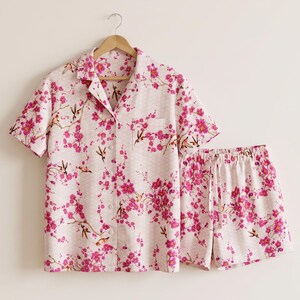 May include: A short-sleeve button-down pajama shirt and matching elastic-waist shorts set in a light pink fabric. The garment features a repeating pattern of vibrant magenta cherry blossoms and small brown hummingbirds on a subtle diamond grid.
