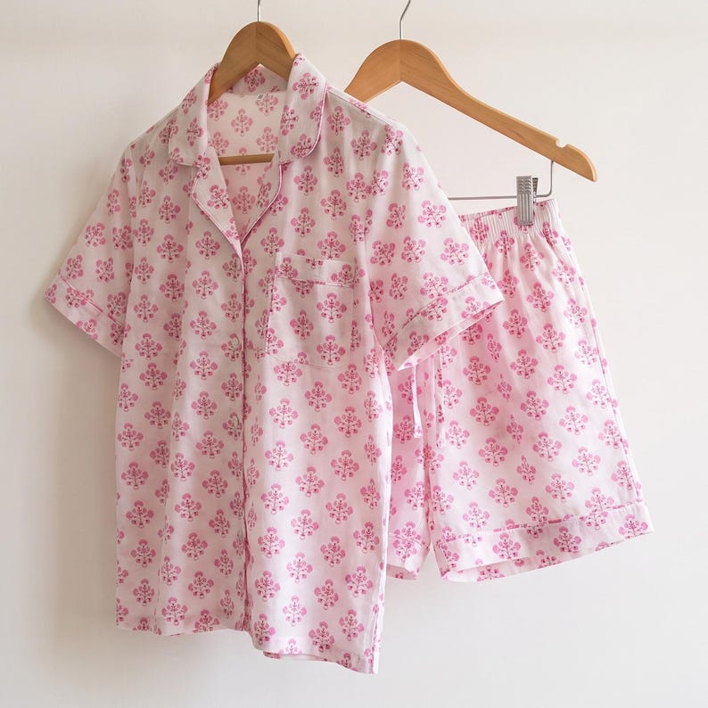 May include: A two-piece pajama set consisting of a short-sleeved button-down shirt with a collar and a chest pocket, and matching shorts with an elastic waistband. Both pieces are white with a repeating pink floral block print pattern.