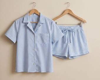 May include: A short-sleeved button-down shirt and matching shorts set featuring a vertical light blue and white striped pattern. The shirt includes a notched collar and a single chest pocket, while the shorts have an elastic waistband with a drawstring.