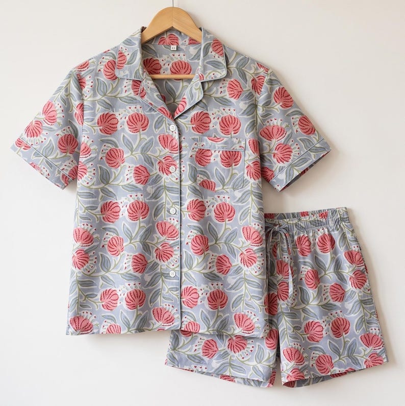 May include: A short-sleeve button-down shirt and matching shorts set in a light blue fabric featuring a repeating pattern of pink floral blossoms with green leaves. The shirt includes a notched collar, a single chest pocket, and white buttons.