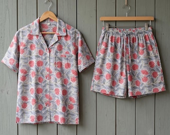 May include: A short-sleeved button-down shirt and matching shorts set featuring a repeating pattern of red flowers and grey leaves on a light grey background. The shirt includes a notched collar and a single chest pocket, while the shorts have an elasticized waistband with a drawstring.