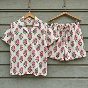 May include: A short-sleeved button-down shirt and matching shorts set in an off-white fabric featuring a repeating pattern of pink flowers with green stems and leaves. The shirt has a notched collar and a chest pocket, while the shorts have a drawstring.