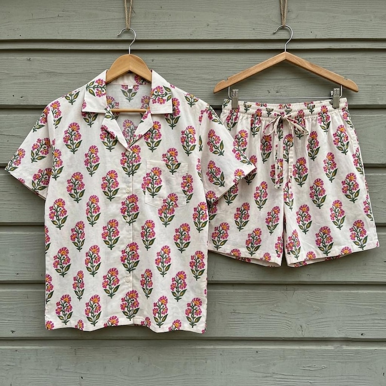 May include: A short-sleeved button-down shirt and matching shorts set in an off-white fabric featuring a repeating pattern of pink flowers with green stems and leaves. The shirt has a notched collar and a chest pocket, while the shorts have a drawstring.