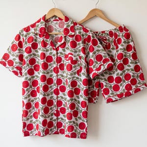 May include: A short-sleeve button-down pajama shirt and matching shorts set featuring a white base with a repeating pattern of red circular flowers and green leaves. The shirt includes a notched collar, a single chest pocket, and red piping.