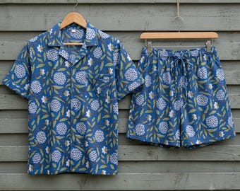 May include: A short-sleeve button-down shirt and matching shorts set in a vibrant blue fabric featuring a repeating pattern of white floral clusters and green leaves. The shirt includes a notched collar and a single chest pocket, while the shorts feature an elasticized waistband with a drawstring tie.