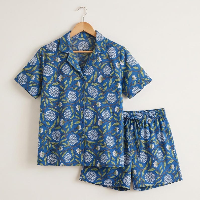 May include: A short-sleeved button-down shirt and matching shorts set in a vibrant blue color. The fabric features a repeating pattern of white hydrangea-like flower clusters and green leaves with brown stems, creating a botanical print.
