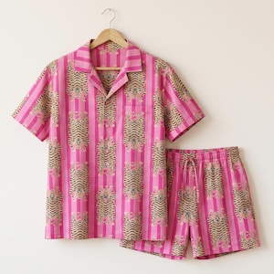 May include: A short-sleeved button-down shirt and matching shorts set featuring a repeating pattern of tigers on a background of vertical pink and light pink stripes. The shirt has a notched collar, a front pocket, and a button-front closure.