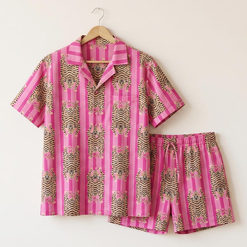 May include: A short-sleeved button-down shirt and matching shorts set featuring a repeating pattern of tigers on a background of vertical pink and light pink stripes. The shirt has a notched collar, a front pocket, and a button-front closure.