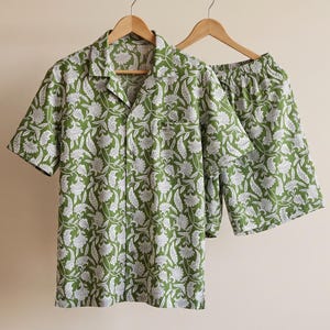 May include: A short-sleeve button-down shirt and matching shorts set in a vibrant olive green color. The fabric features a repeating white floral and paisley pattern throughout. The shirt has a notched collar and a single chest pocket.