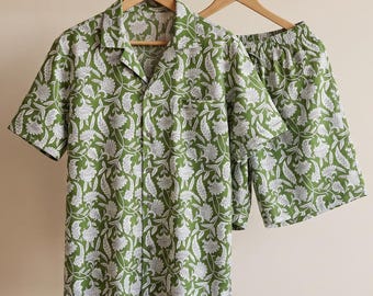 May include: A short-sleeve button-down shirt and matching shorts set in a vibrant olive green color. The fabric features a repeating white floral and paisley pattern throughout. The shirt has a notched collar and a single chest pocket.