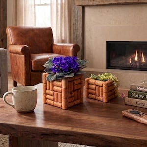 May include: Two square planters constructed from stacked natural corks, one containing a purple African violet and the other a small succulent. A speckled cream ceramic mug and a wooden remote control sit nearby on a dark wood table.