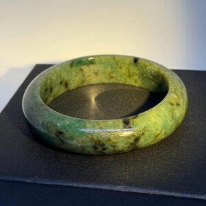 May include: A smooth, circular bangle bracelet crafted from polished green stone. The surface features a mottled appearance with varying shades of vibrant emerald and lime green, interspersed with dark black inclusions and natural mineral veining.