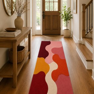 May include: A rectangular runner rug featuring a vibrant, abstract pattern of interlocking organic shapes in shades of deep burgundy, bright red, orange, soft pink, and cream, laid out on a wooden floor in a hallway setting.