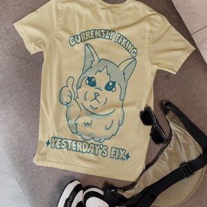 Puede incluir: Una camiseta de color amarillo claro con un gráfico azul oscuro de un gato haciendo un gesto de pulgar hacia arriba. El texto sobre el gato dice CURRENTLY FIXING y el texto debajo dice YESTERDAY'S FIX. El diseño incluye pequeñas estrellas a los lados.