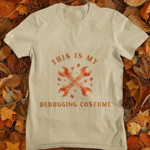 May include: A beige short-sleeved t-shirt featuring the text "THIS IS MY DEBUGGING COSTUME" in brown letters. The center graphic displays two crossed wrenches filled with an orange and brown autumn leaf pattern, surrounded by small stars and acorns.