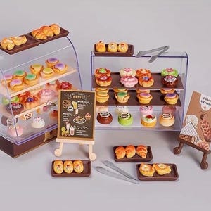 Op de afbeelding: Een miniatuur bakkerijset met twee doorzichtige acryl vitrines gevuld met diverse kleine gebakjes, twee houten ezels met een menukaart en een bordje met de tekst Baked & Frosted, diverse bruine dienbladen en twee zilverkleurige metalen tangen.
