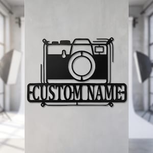 May include: A black metal wall sign featuring a stylized camera outline positioned above a rectangular nameplate with the text CUSTOM NAME cut out in a bold, sans-serif font. The design is flat, monochromatic, and symmetrical in its layout.