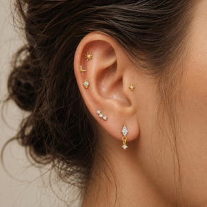 May include: A collection of gold-toned earrings worn in a single ear, featuring a starburst stud, an L-shaped stud, a diamond-shaped stud, a triple-stone stud, a tragus star stud, and a dangling marquise-cut stone earring with a small star accent.