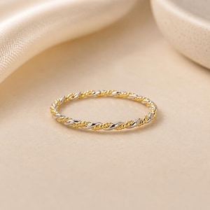 Può includere: Un anello sottile e delicato caratterizzato da un design intrecciato che alterna metallo color argento lucido e metallo color oro testurizzato. La fascia presenta un motivo continuo a corda, creando un aspetto sottile e raffinato per il dito.
