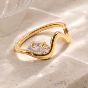 May include: A gold-toned ring featuring a wavy, undulating band design. The ring is set with a single, clear, marquise-cut gemstone held in place by four small prongs, positioned along the curve of the band with a polished, reflective finish.