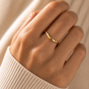 May include: A gold-toned curved ring featuring a wavy band design. The surface of the ring is accented with three small, embedded clear stones arranged in a line along the centre of the band, creating a subtle sparkle on the polished metal finish.